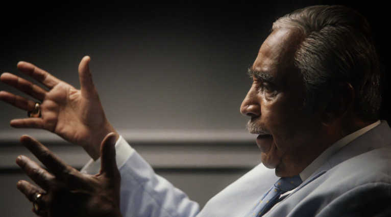 What Is a Talking Head Shot? Definition + How to Use It 3 Side-profile shot of an older man in a light suit speaking with animated hand gestures, lit dramatically against a dark background.