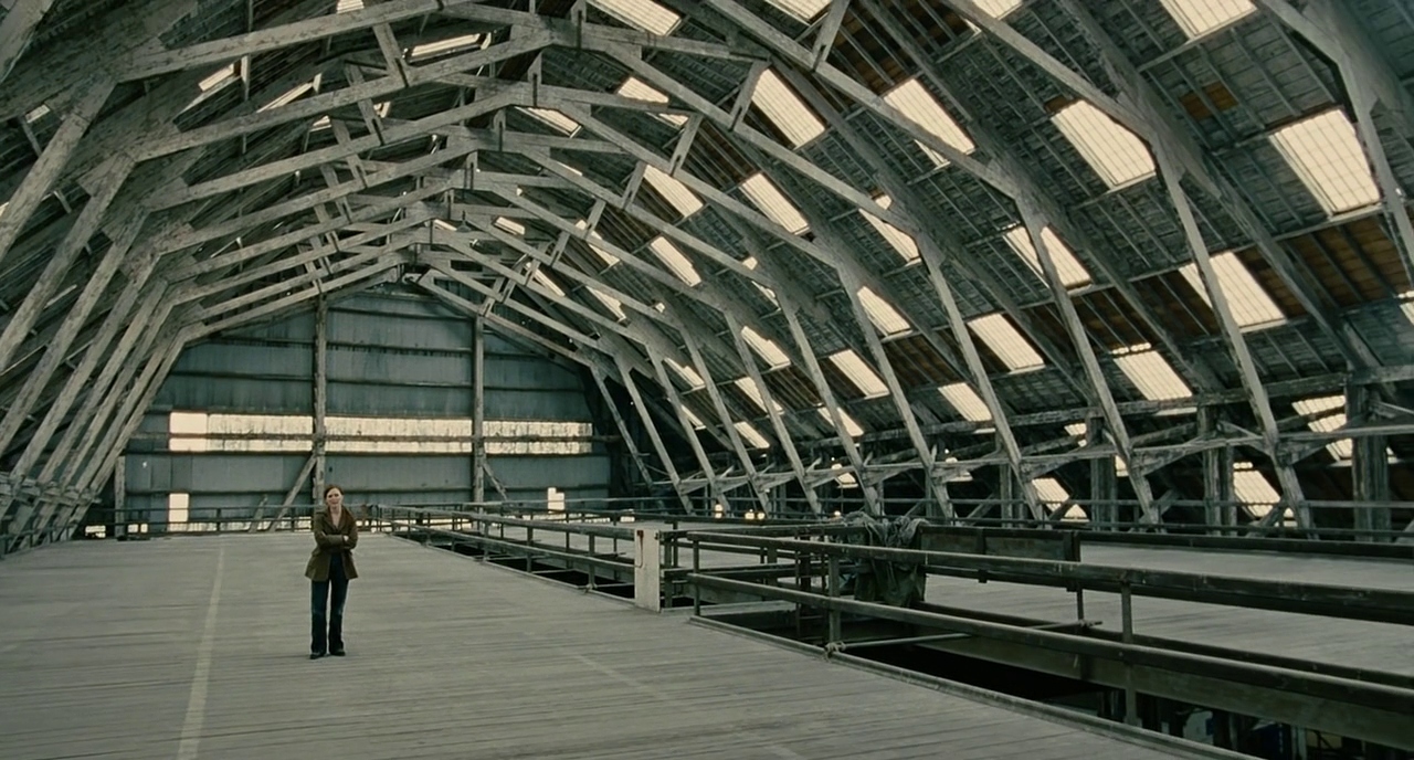 What Is Dystopian Fiction? Meaning + Examples from Film 6 A woman stands alone in a vast, empty warehouse with wooden floors and a high metal roof. The space feels cold and deserted.