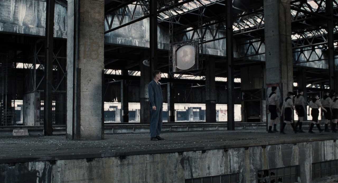 What Is Atmosphere in Writing? Definition & Examples 3 Winston stands in a grey industrial hall under a large surveillance screen while children in uniform march past in the background.
