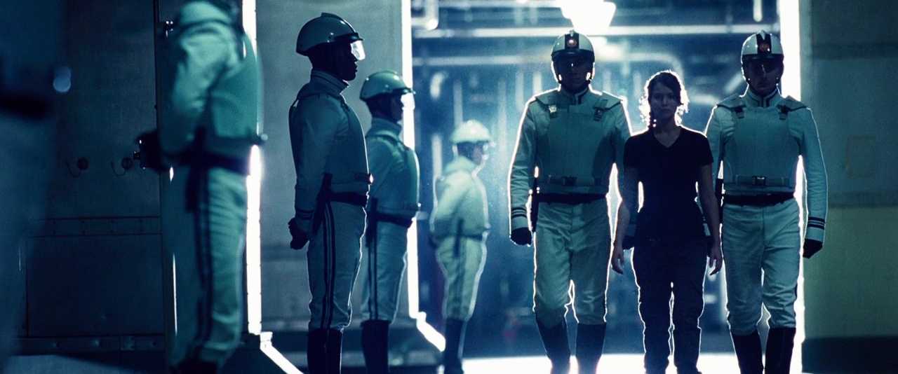 What Is Dystopian Fiction? Meaning + Examples from Film 4 Katniss walks between two Peacekeepers in white uniforms as others stand guard in formation. She is framed by bright light at the end of a dark industrial hallway.