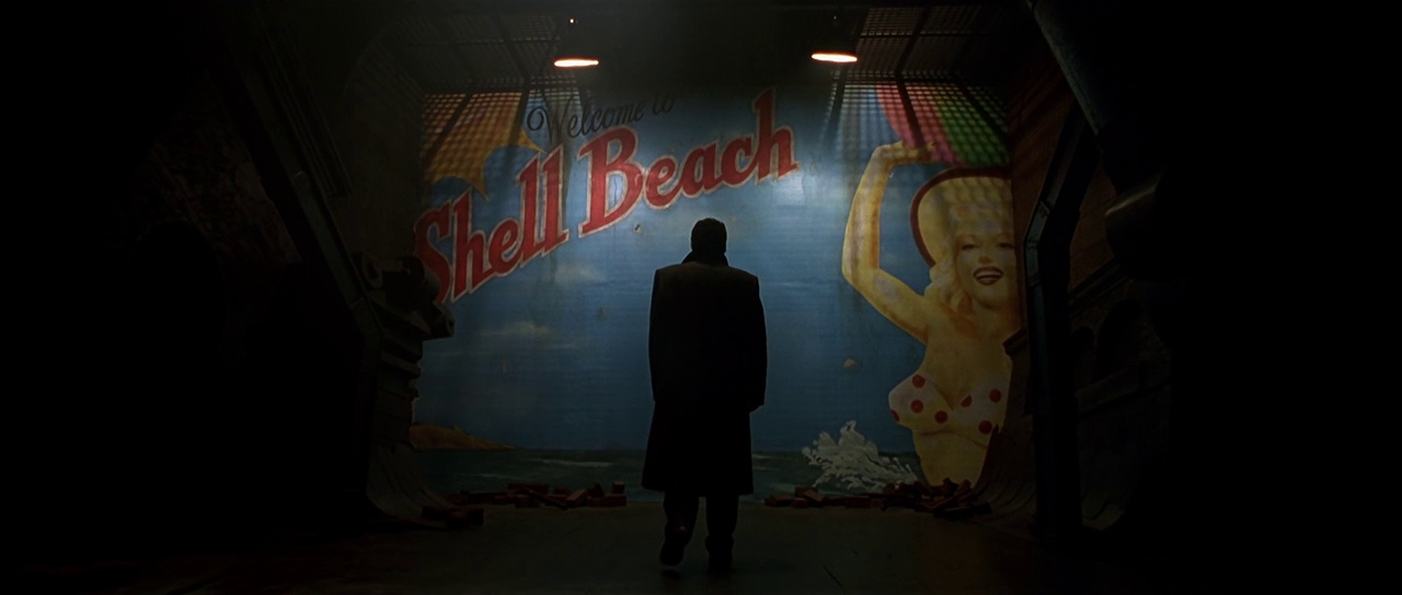 What is Plato's Allegory of the Cave? Meaning, Films & Philosophy Explained 5 A man in a dark coat stands in front of a painted wall showing “Welcome to Shell Beach,” revealing the illusion of a fake destination