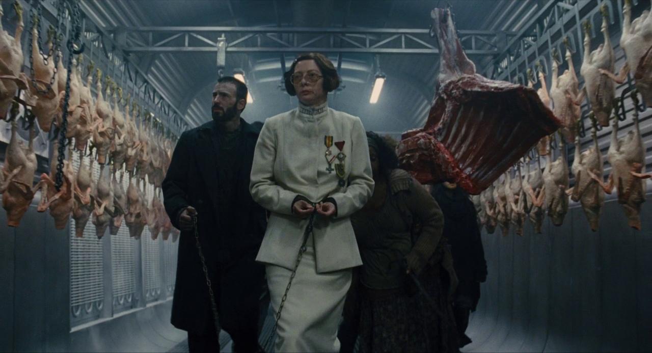 What Is Dystopian Fiction? Meaning + Examples from Film 2 A woman in a white uniform walks through a meat storage car, flanked by prisoners in chains. Sides of beef and rows of hanging chickens line the walls.
