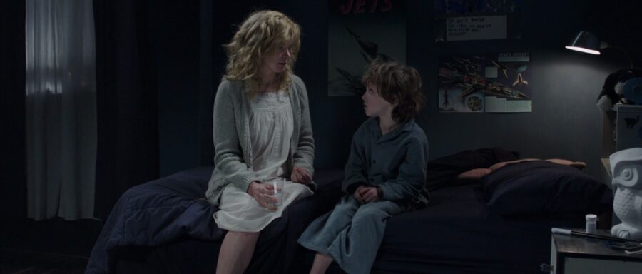 What Is Inductive Reasoning? How It Works in Film and Storytelling 2 A woman in a white nightgown and gray cardigan sits on the edge of a dark bed, talking to a young boy in pajamas. The dimly lit room has dark walls with space posters and a desk lamp casting soft light. The boy looks up at the woman with concern.