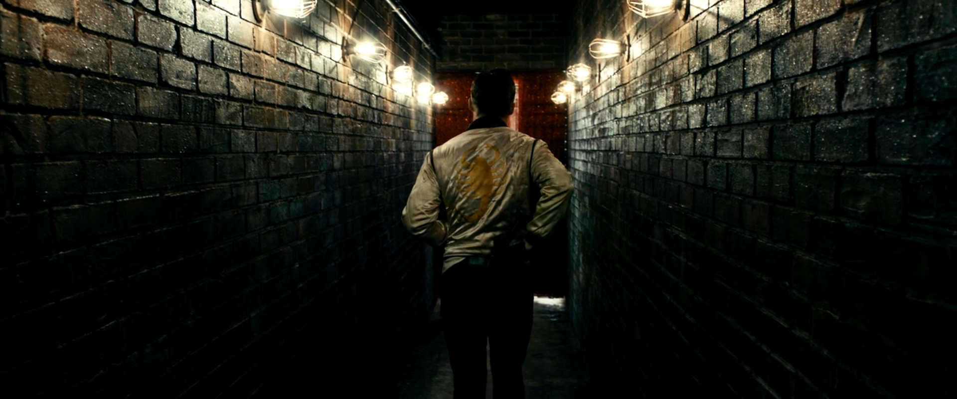 The Scorpion & the Frog Story Explained 2 The Driver walks down a dark brick hallway, wearing a jacket with a large golden scorpion on the back, lit by overhead lights.