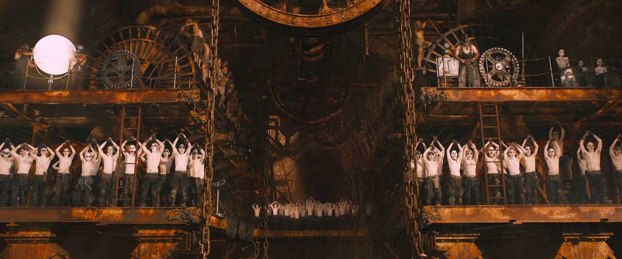What Is World Building in Movies? Definition, Meaning & Examples 10 Rows of pale, shirtless workers stand on rusted platforms beneath giant gears, raising their arms in unison inside an industrial cavern.