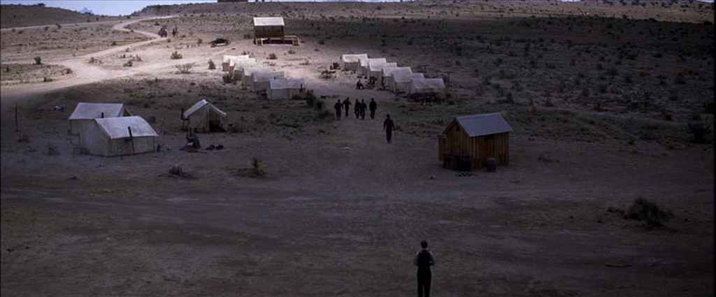 Atmosphere in Film: Definition, Meaning & Examples 3 Man walks toward a group of tents and wooden shacks in a barren desert landscape