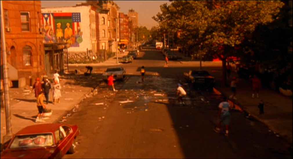What Is World Building in Movies? Definition, Meaning & Examples 7 A wide shot of a Brooklyn street with people gathering, trash scattered, and a large mural on the side of a brick building under intense sunlight.