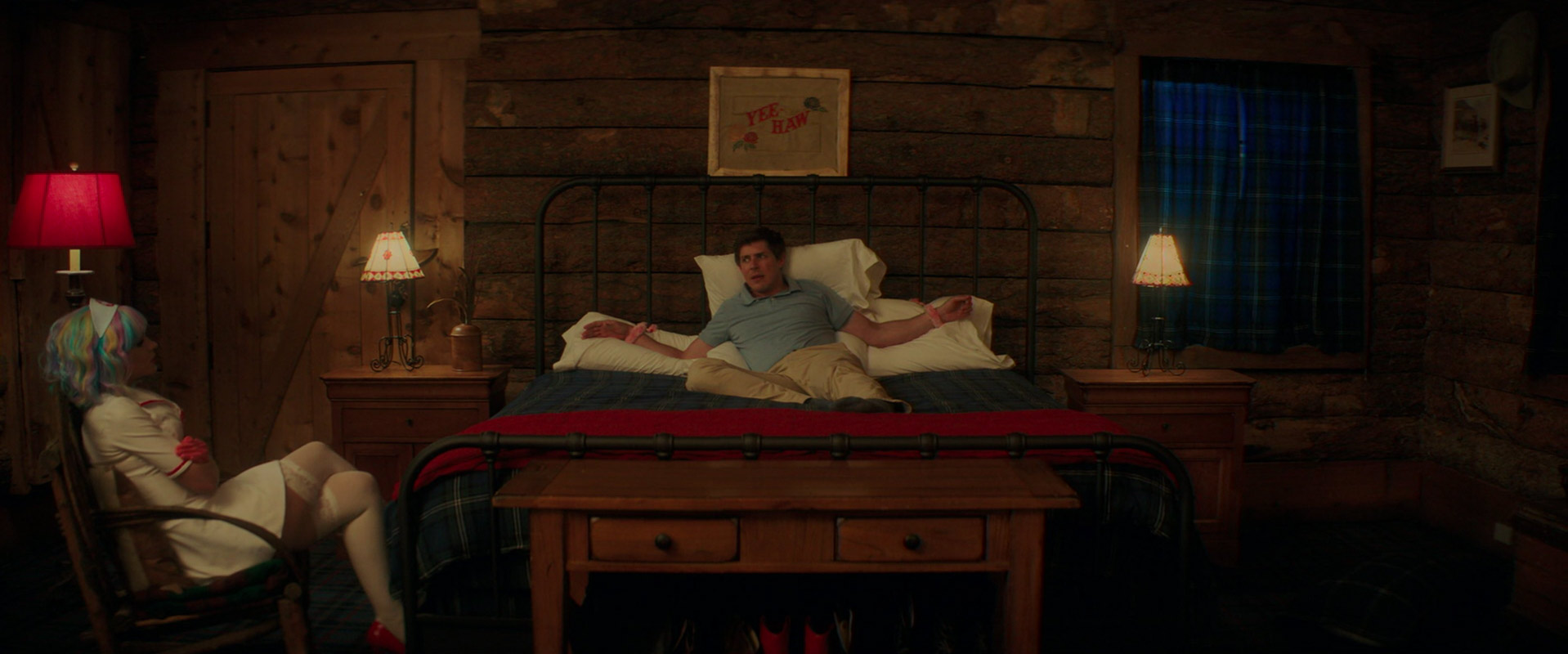 What Is the Male Gaze in Film? Definition +How Movies Use It 8 A man lies on a bed with his wrists tied while a woman in a nurse outfit and rainbow wig sits in a chair, facing him in a log cabin room.