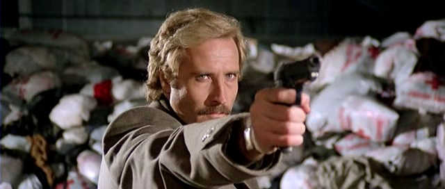 What Is Italian Exploitation Cinema? From Sex to Cannibalism 4 A man in a suit points a handgun with both hands inside a warehouse full of plastic-wrapped packages