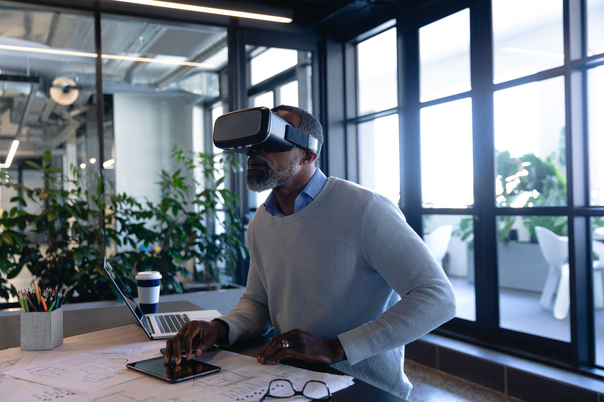 What Is Virtual Reality? Definition, Film Uses & Future Trends 5 Man using VR headset at desk with tablet, laptop, and design plans