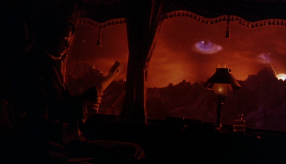 What Is Motivated Lighting? Definition, Why, and How It’s Used on Set 6 Dark interior with a seated figure in silhouette at left, a lit table lamp on a desk near center, and a bright orange window view with clouds and faint eyes in the sky.