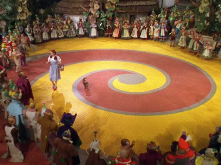 What Is Technicolor? Definition, Technology, and Film Examples 6 A wide overhead shot of Dorothy on a large yellow-and-red spiral-patterned plaza, surrounded by a ring of people in colorful costumes.