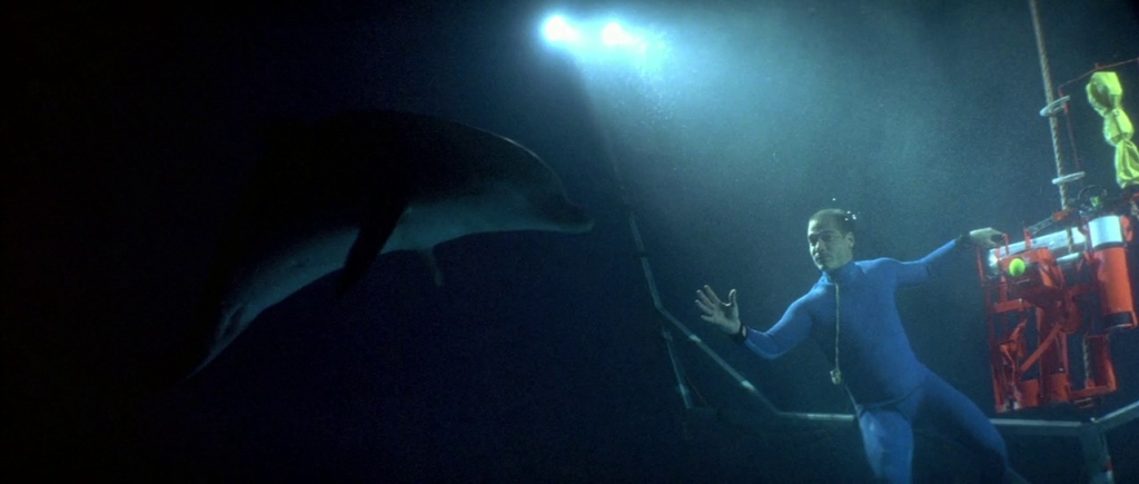 What Is Motivated Lighting? Definition, Why, and How It’s Used on Set 5 Underwater shot of a diver in a blue suit reaching out beside a red rig, with bright dive lights shining from above and a shark in silhouette on the left.