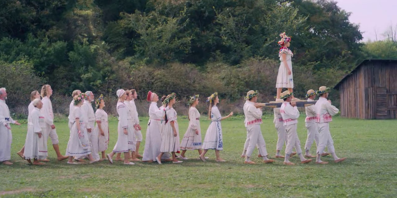 What is a 2:1 Aspect Ratio? Definition, Workflow & Film Examples 2 A line of barefoot villagers in white outfits walks across a grassy field. Several people carry a wooden platform with a flower-crowned woman standing on it. A small wooden building sits on the right, with dense trees in the background.