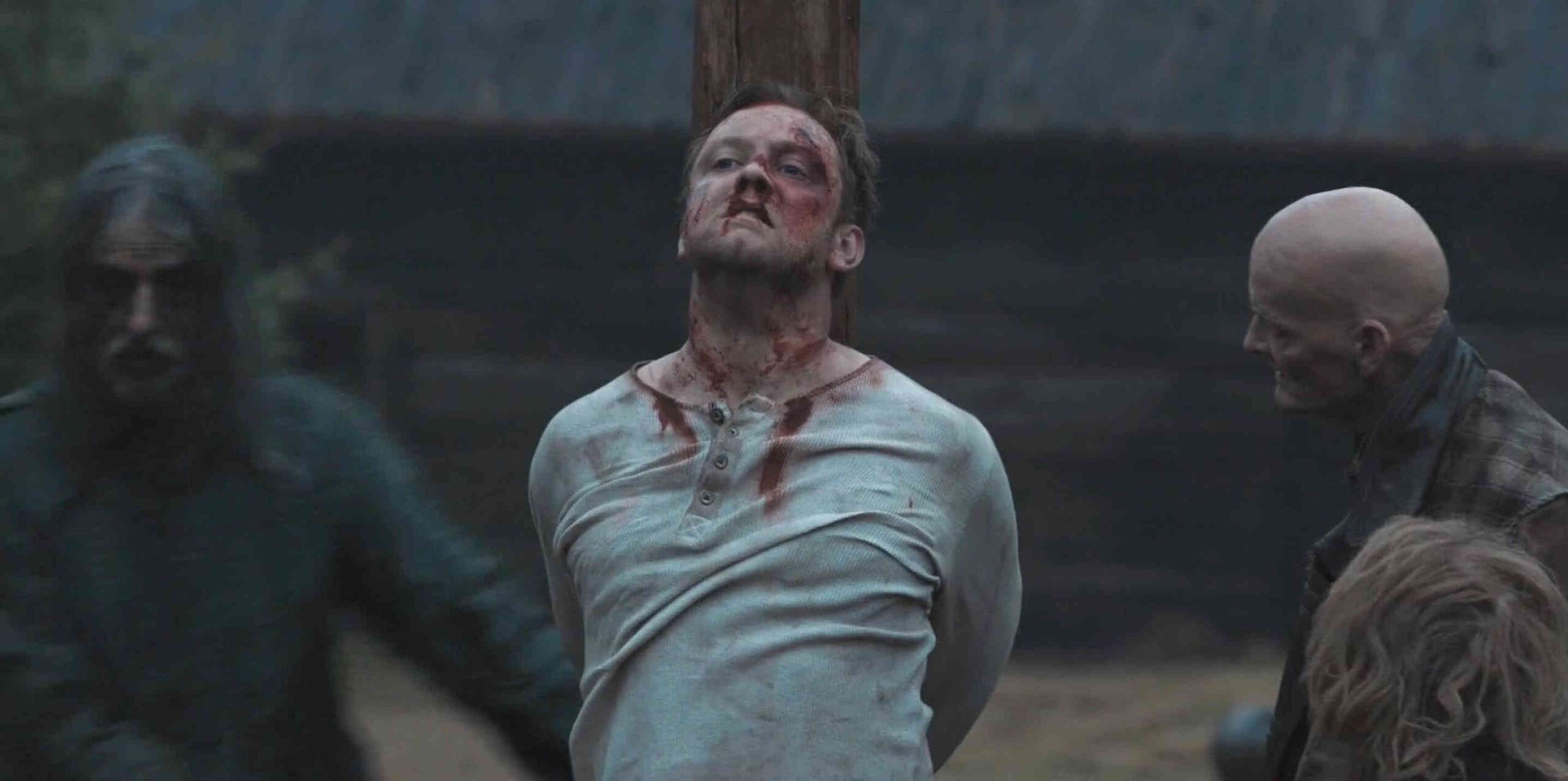 What is a 2:1 Aspect Ratio? Definition, Workflow & Film Examples 5 A bruised, bloodied man in a light shirt is tied upright against a wooden post. A bald man leans in from the right, and shadowy figures appear near the left and lower-right edges in a dark outdoor setting.