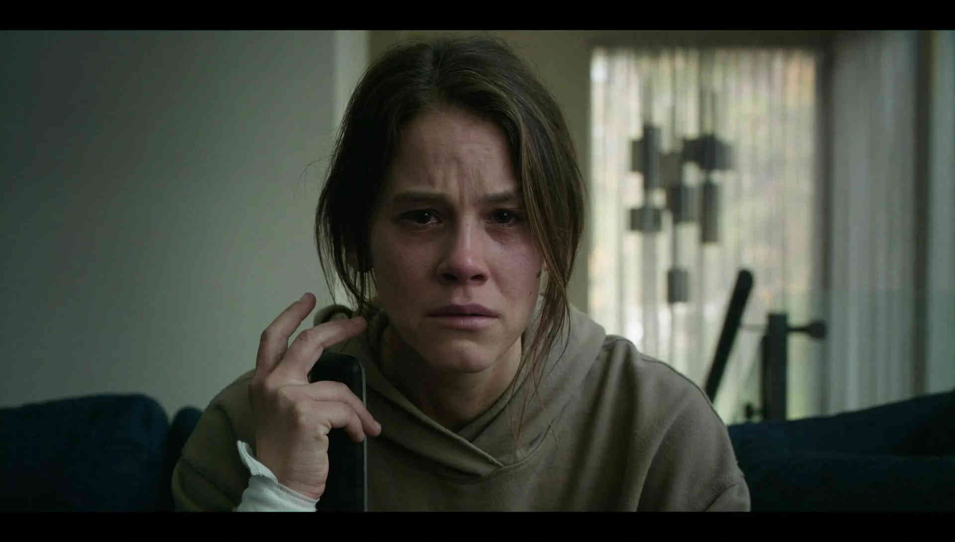 What is a 2:1 Aspect Ratio? Definition, Workflow & Film Examples 6 A close shot of a worried woman in a hoodie holding a phone receiver near her face while sitting on a dark couch. A bright window with patterned curtains and exercise equipment sit in the background, with black letterbox bars.