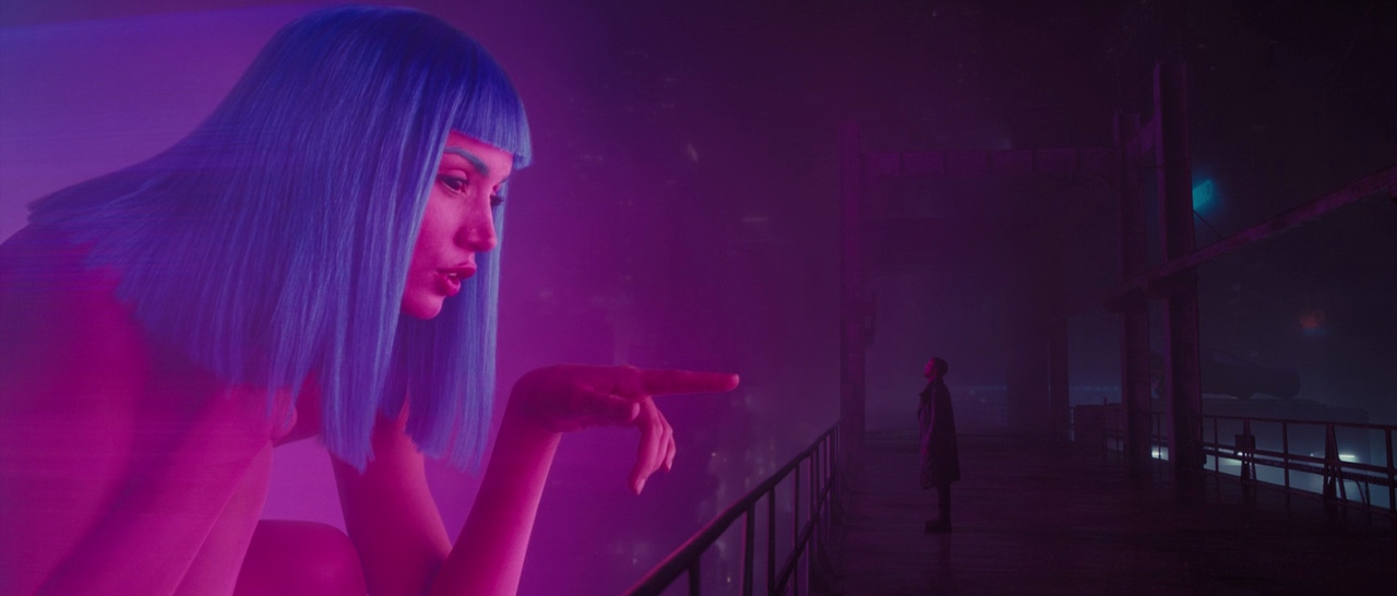 What Is Motivated Lighting? Definition, Why, and How It’s Used on Set 2 A giant pink-purple holographic woman with blue hair leans in from the left and points toward a lone man standing on a dark balcony, surrounded by foggy neon light and a dim futuristic city.