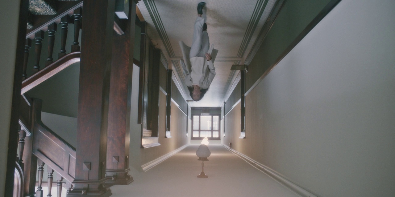 What is a 2:1 Aspect Ratio? Definition, Workflow & Film Examples 3 An upside-down shot of a long hallway with stairs and dark wood railings on the left. A person appears “standing” on the ceiling near the far end of the hall, with a lamp and a bright doorway in the distance.