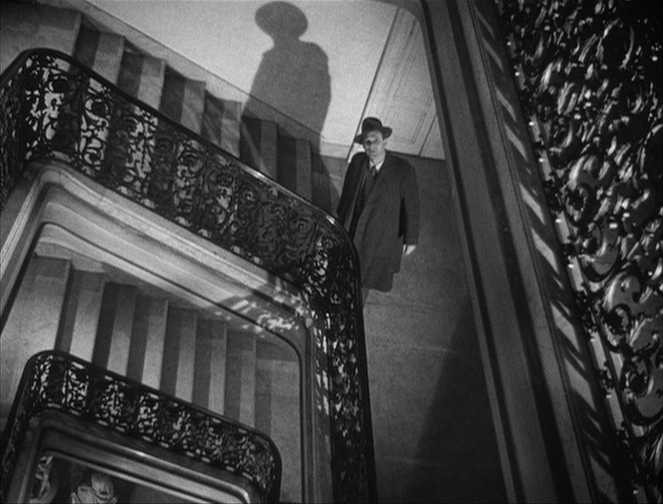 Best Mystery Movies of All Time: 25 Picks Explained 11 Black-and-white shot looking up a stairwell with ornate railings; a man in a coat and hat stands on an upper landing, casting a large shadow on the wall.