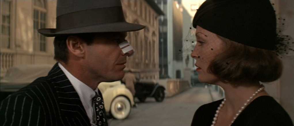 Best Mystery Movies of All Time: 25 Picks Explained 12 A man in a fedora with a bandage on his nose faces a woman wearing a dark hat and pearls on a sunlit street, with a vintage car in the background.