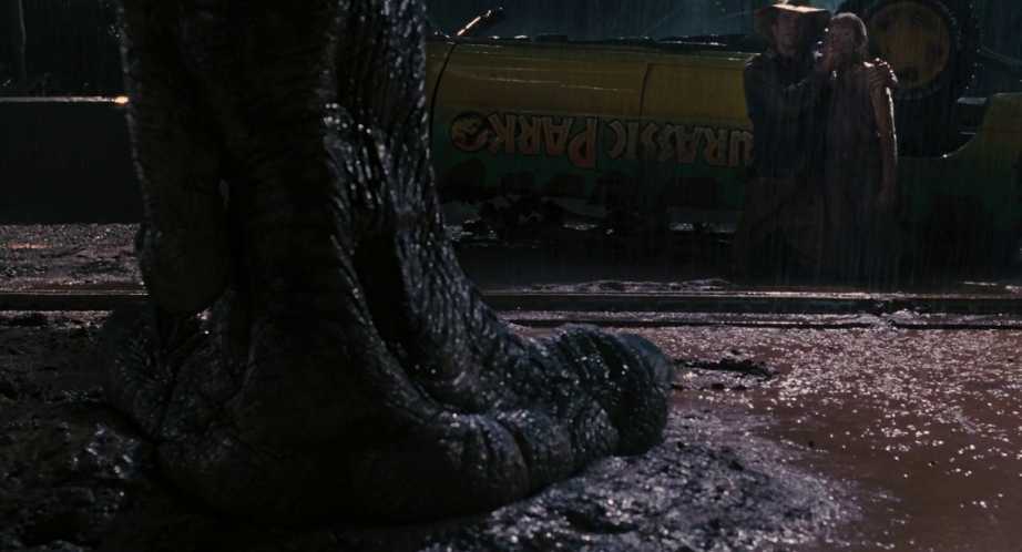 T. rex breakout at night in Jurassic Park The foot of a Tyrannosaurus rex lands in the mud at night while two characters stand frozen beside an overturned Jurassic Park vehicle in heavy rain.
