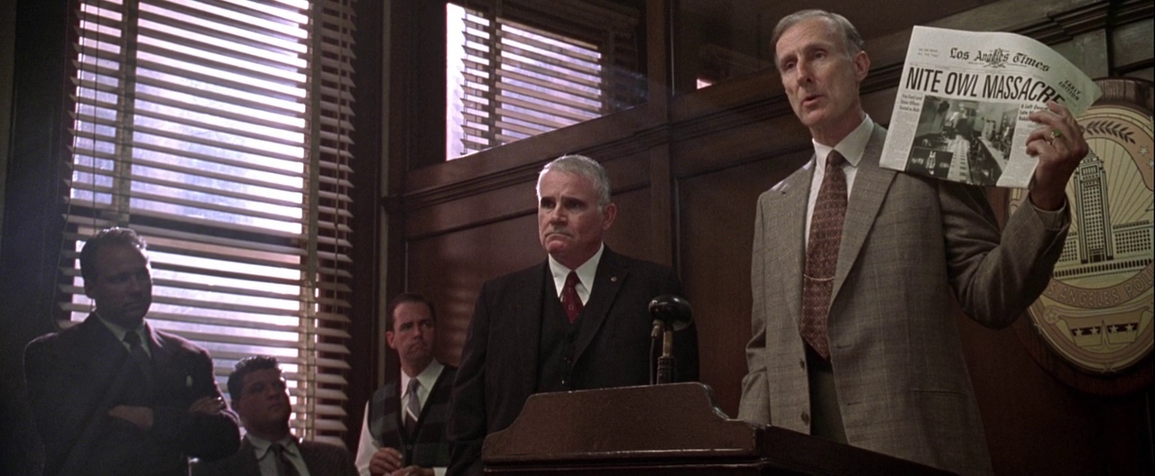 Best Mystery Movies of All Time: 25 Picks Explained 13 Several men stand in a police briefing room; one man at a podium holds up a newspaper with the headline “Nite Owl Massacre,” with venetian blinds casting light across the room.
