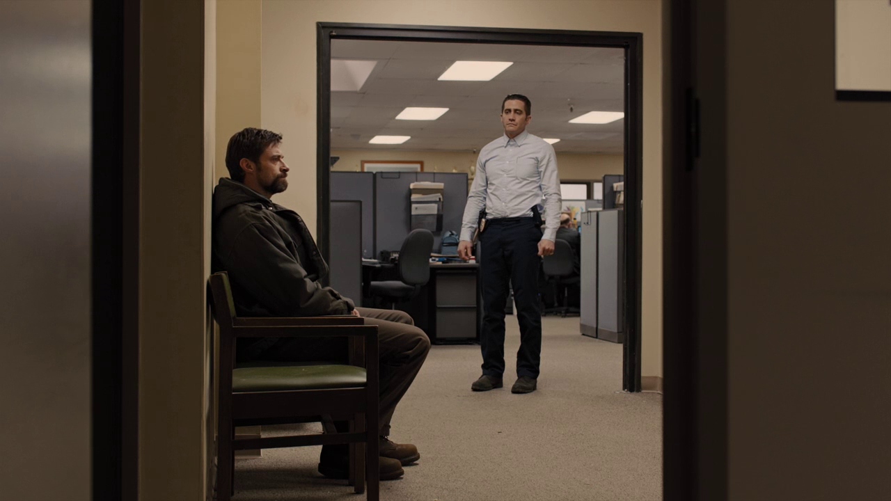 Best Mystery Movies of All Time: 25 Picks Explained 25 A man sits in a chair in a hallway while another man stands in a doorway to an office area with cubicles and fluorescent lights.