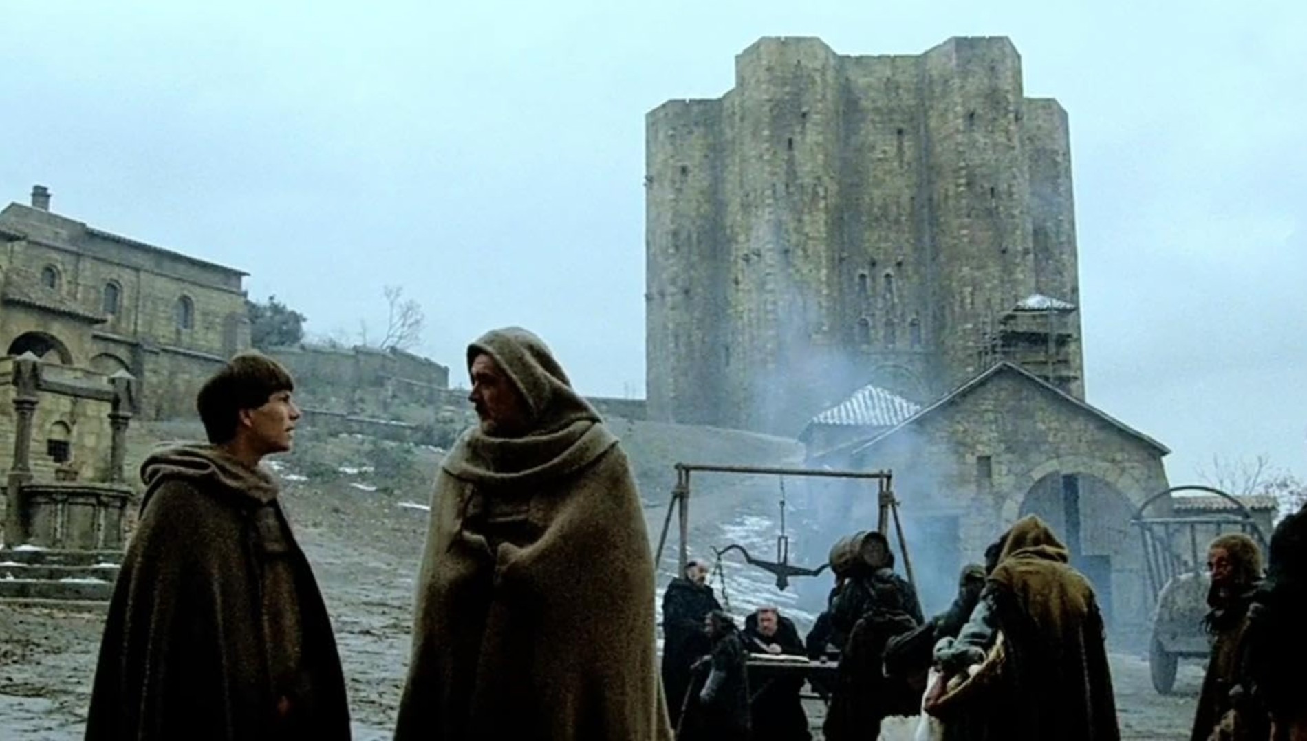 Best Mystery Movies of All Time: 25 Picks Explained 26 Two robed men stand in a cold stone courtyard near an abbey, with a large tower in the background and several figures gathered near smoke and carts.