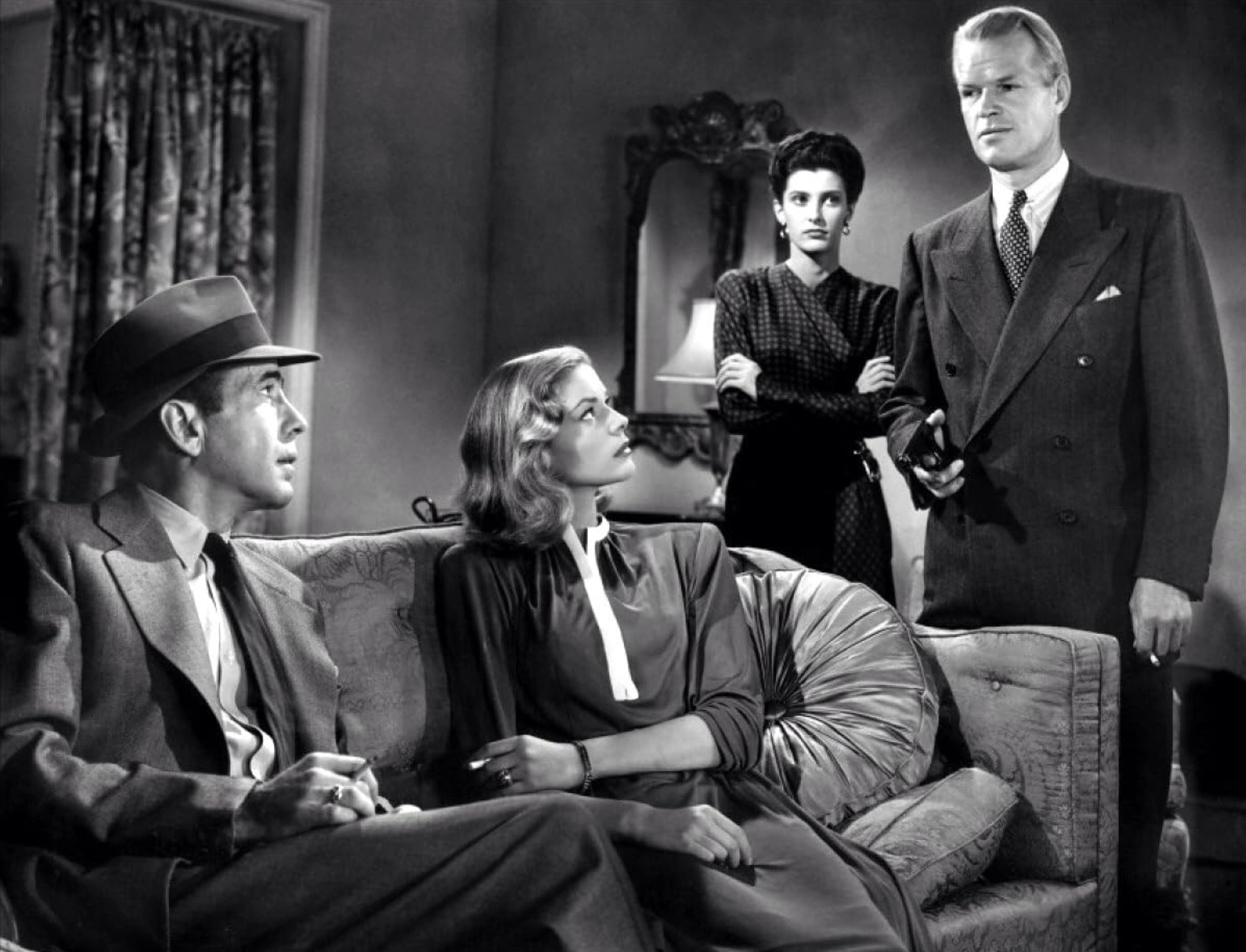 Best Mystery Movies of All Time: 25 Picks Explained 10 Black-and-white scene with a man in a hat seated on a couch beside a woman, while another man stands holding a small object; a woman stands behind them with arms crossed in a living room.