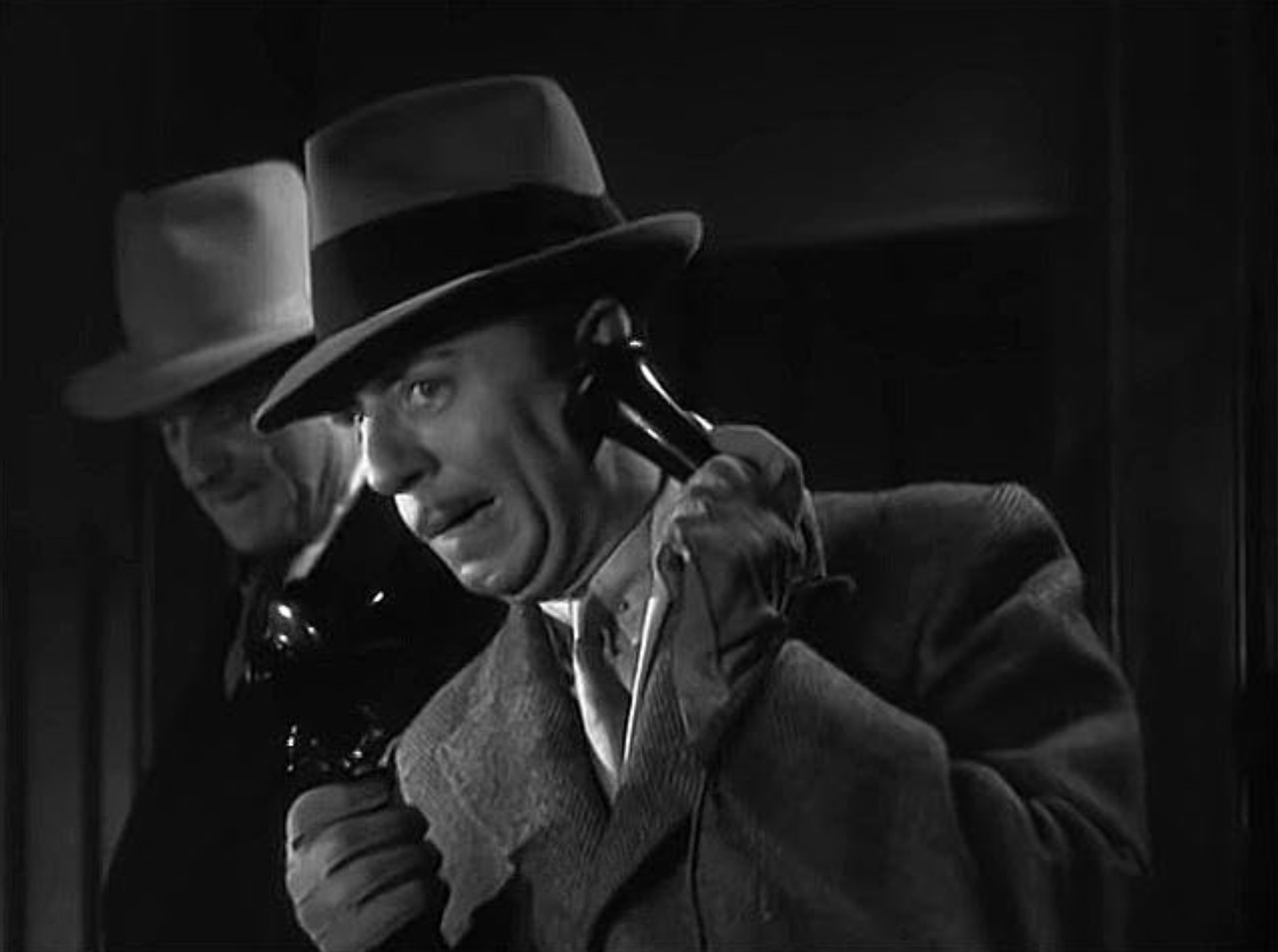 Best Mystery Movies of All Time: 25 Picks Explained 3 Black-and-white close-up of a man in a fedora and coat holding a telephone receiver, with another man in a light hat partly visible behind him in shadow.