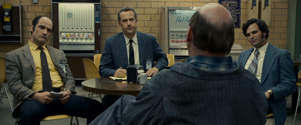 Best Mystery Movies of All Time: 25 Picks Explained 22 Three men sit at a table in a break room facing a man whose back is to the camera; vending machines and brick walls are behind them.
