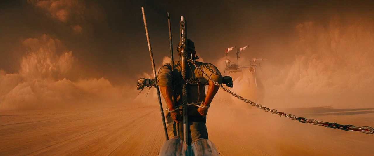 Rhythmic Editing: How Cut Timing Controls the Audience 4 Max Rockatansky is chained to the front of a moving vehicle as it races across a dusty desert road, with another vehicle following behind.
