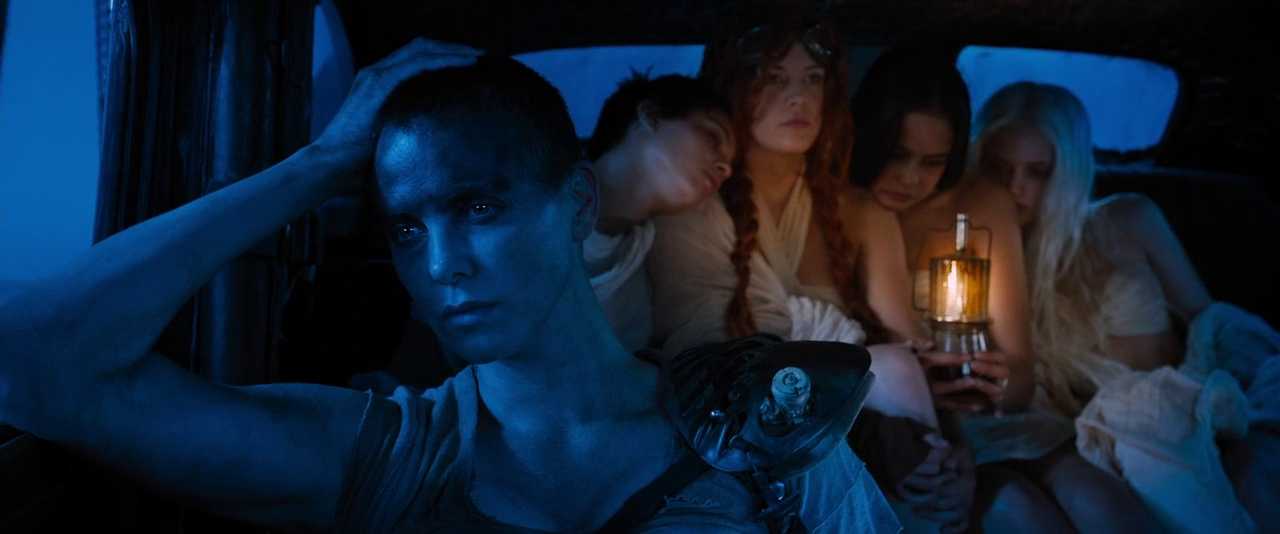 Neoformalism Film Theory: Definition, Method, Examples 4 Furiosa sits in the front of a dark vehicle interior under blue light, while the Wives huddle behind her and one holds a lit lantern.