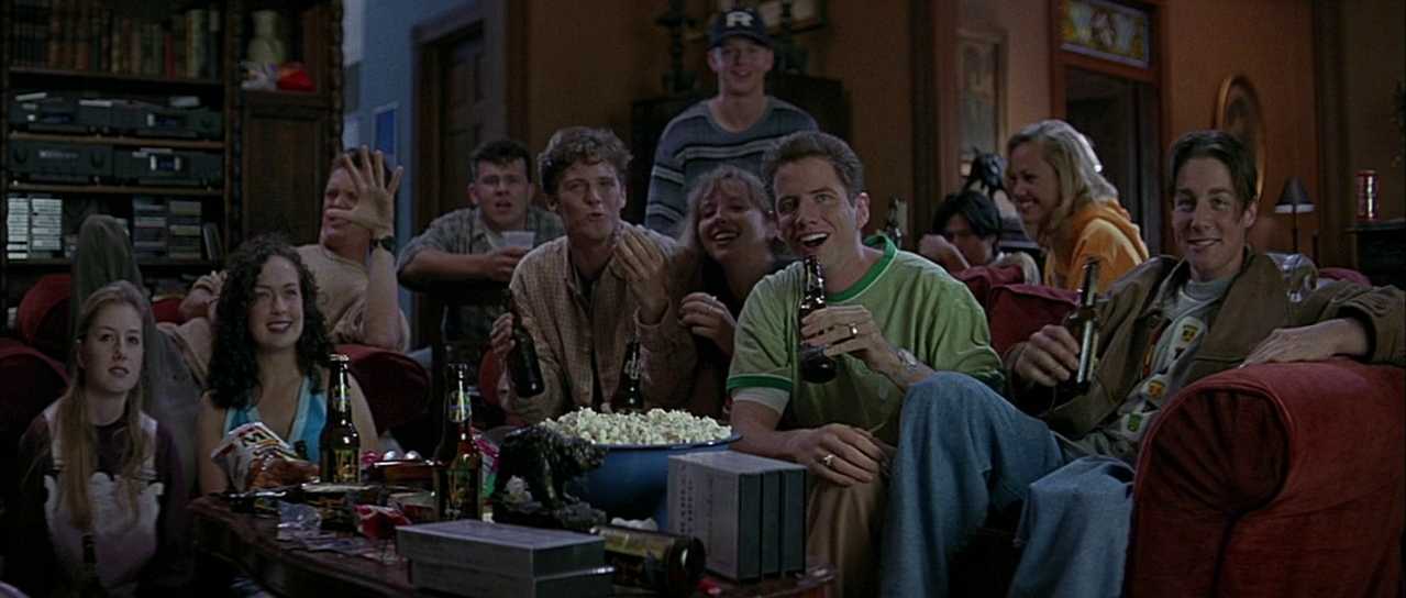 Postmodern Film Theory: What It Is and How It Works 3 A group of teens and young adults sit on couches with beers and a bowl of popcorn, smiling and gesturing toward a TV off screen in a crowded living room.