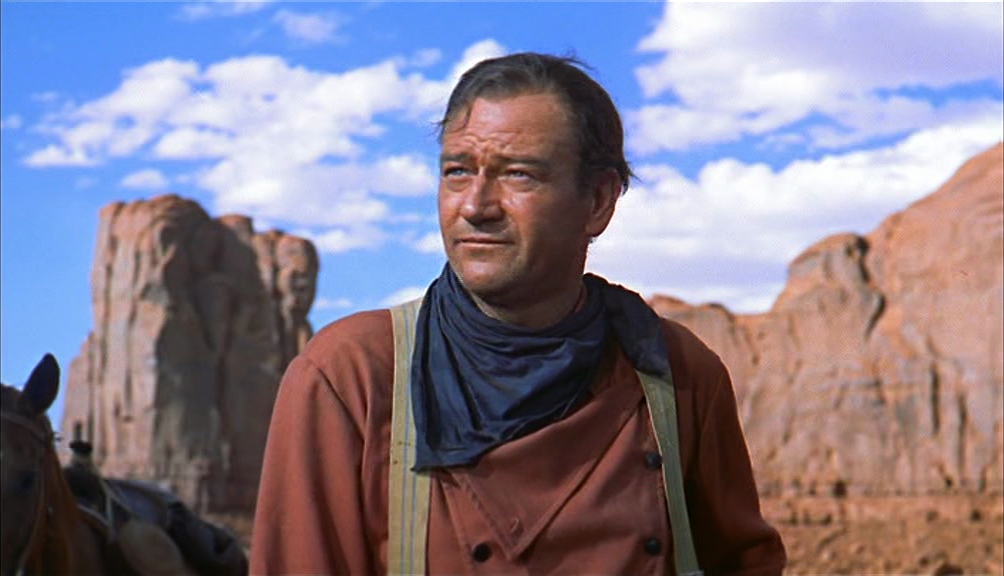 Structuralism in Film Theory: Key Concepts and Analysis 2 A man in a reddish shirt and dark neckerchief stands outdoors in a desert landscape with large red rock formations and a blue sky behind him.