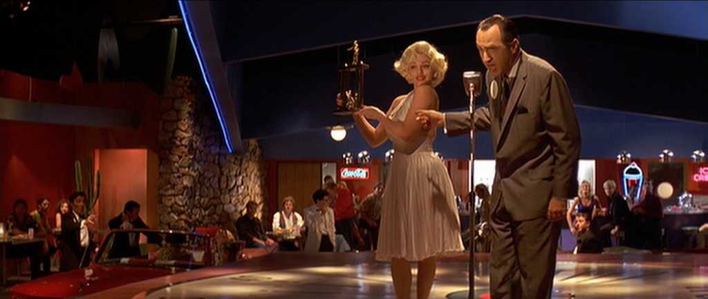 Postmodern Film Theory: What It Is and How It Works 2 A blonde Marilyn Monroe look-alike in a white dress holds a trophy on a small stage while a man in a suit speaks into a microphone, with diners and neon signs in a retro-themed restaurant behind them.