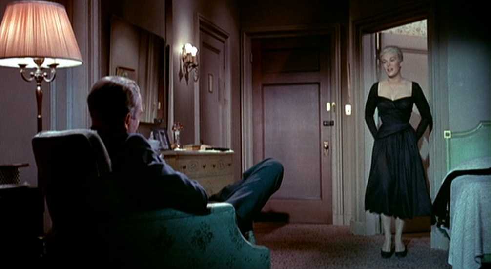 Psychoanalytic Film Theory: Definition and Key Concepts 2 Scottie sits in an armchair in a hotel room facing Judy, who stands near the doorway in a dark dress, with a lamp and bed visible in the room.