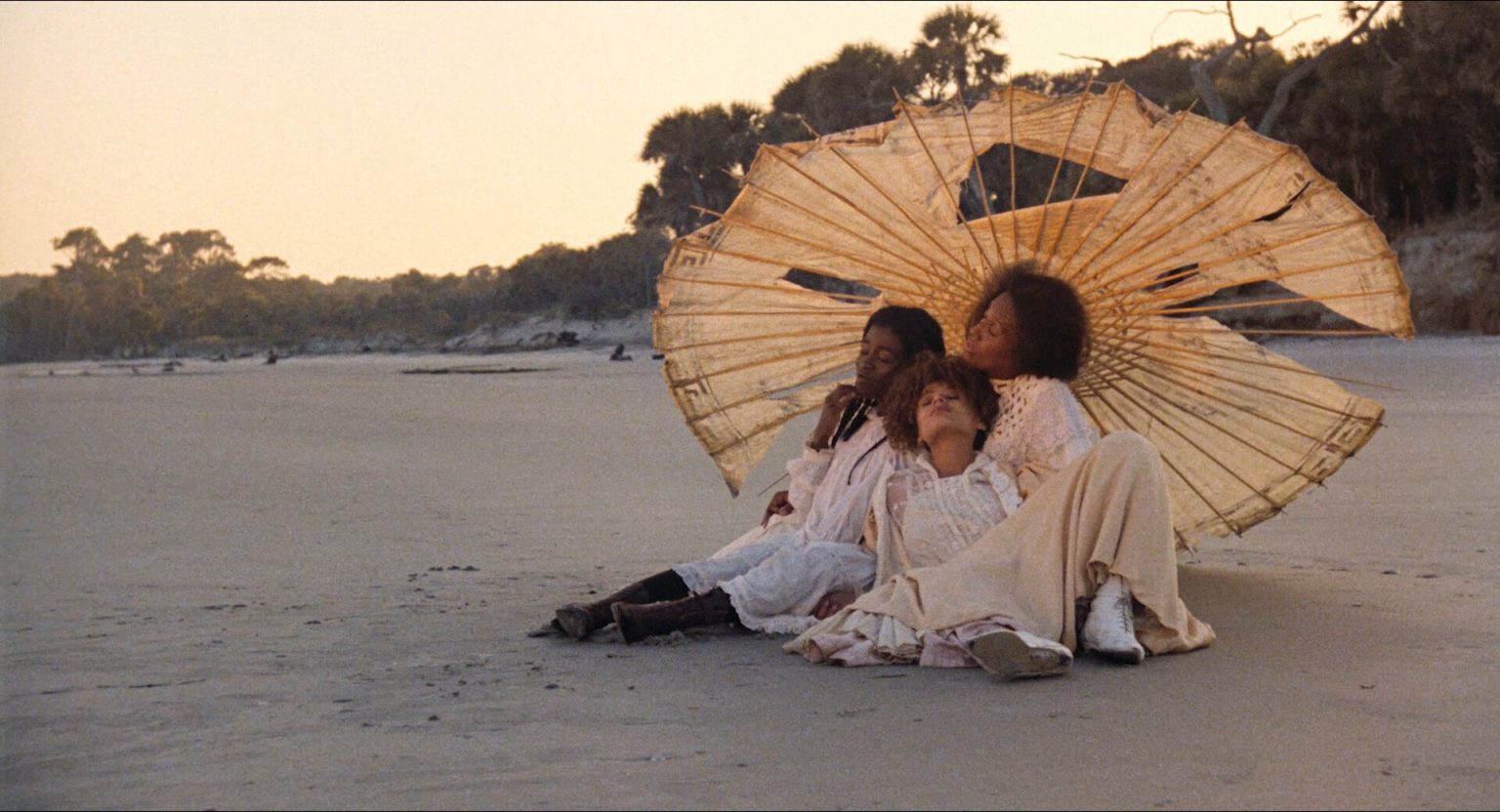 Feminist Film Theory: Concepts, Theorists, and Film Analysis 3 Three women sit close together on a sandy beach beneath a large tattered parasol in Daughters of the Dust (1991), dressed in white Victorian-era clothing in warm evening light.