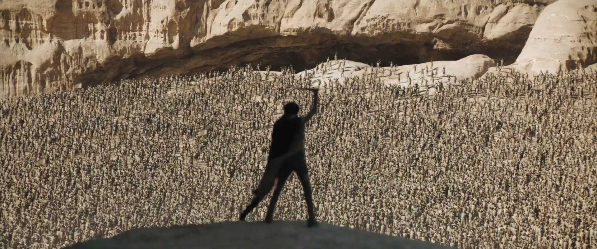 Postcolonial Film Theory: Key Concepts and Analysis 4 Silhouetted Paul Atreides raises an arm on a dune ridge as thousands of Fremen fill the desert basin below, with pale rock cliffs behind them.
