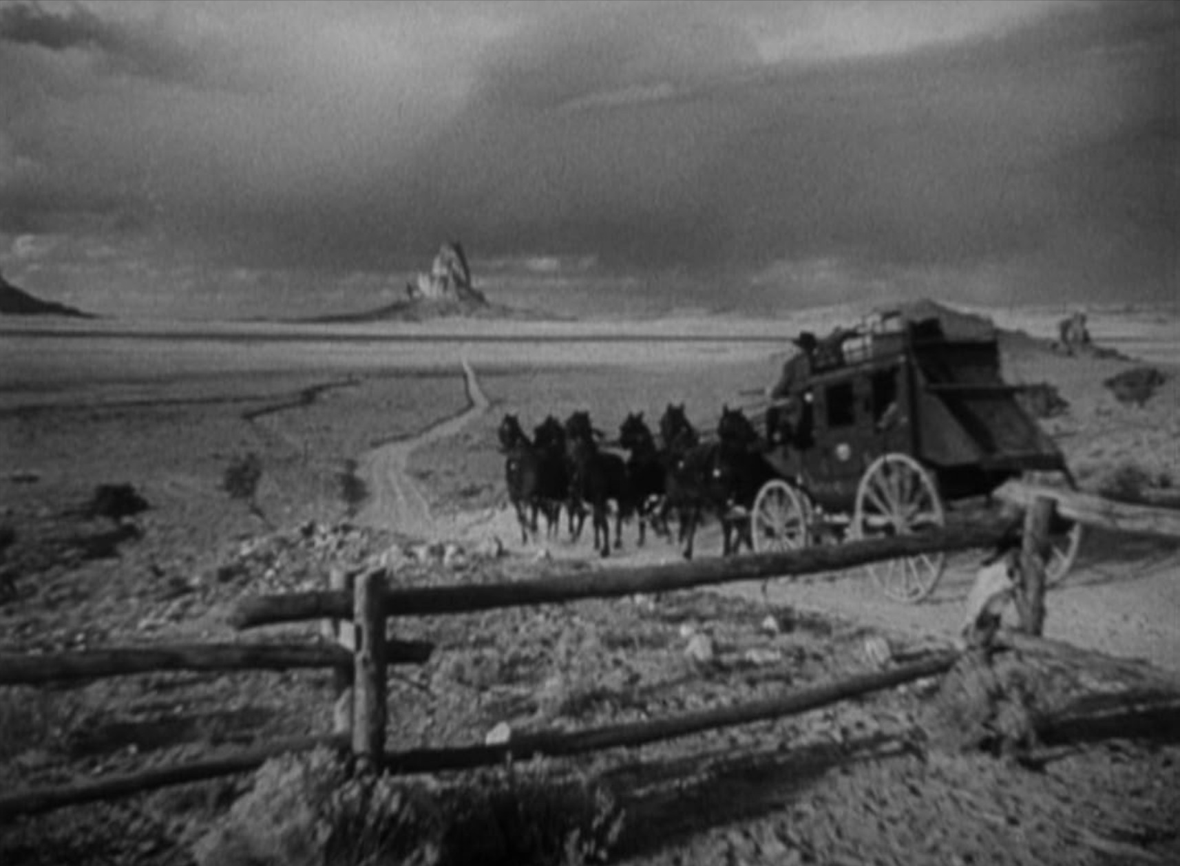 Structuralism in Film Theory: Key Concepts and Analysis 6 Black-and-white wide shot of a stagecoach pulled by a team of horses on a dirt road across a vast plain. A wooden fence runs across the foreground, with distant rock formations under a cloudy sky.